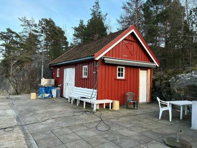 Fotografi av en rød, trepanelert hytte eller uthus med hvite rammer rundt vinduer og dører, plassert på en betongplateform med en bakgrunn av skog og fjell.