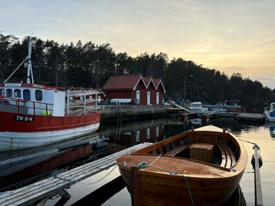 Fotografi av en havn med båter, en kai og røde båthus i bakgrunnen.