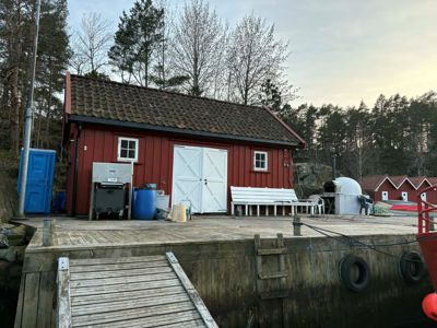 Fotografi av en rød skurbygning med hvite dører og vinduer, plassert på en kai eller holme med skog i bakgrunnen.