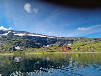 Fotografi av en bygning ved en vannflate med fjell i bakgrunnen. Bildet er merket med en rosa sirkel rundt en spesifikk bygning.