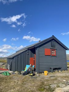 Fotografi av en mørk, lang bygning (trollhytte) med en mann som sitter i en campingstol utenfor.