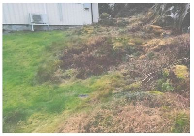 Fotografi av en bygning sett fra siden, med en grønn gressbeite i forgrunnen og en skråning med vegetasjon bak.