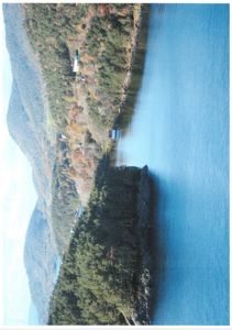 Fotografi av et kystlandskap med fjell, skog og vann. Det er synlig en bygning på kanten av landmassen.