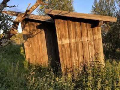 Fotografi av en liten, gammel trebygning (muligens utedo eller utedekk) i en grønn, naturlig omgivelser.