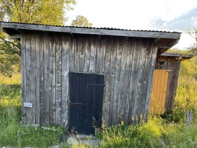 Fotografi av en gammel, trepanelert uthusbygning med mørk dør og tak, omgitt av gress og trær.