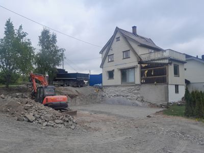 Fotografi av en byggeplass med en delvis ombygd eller renovert hus, en graver og en lastebil.