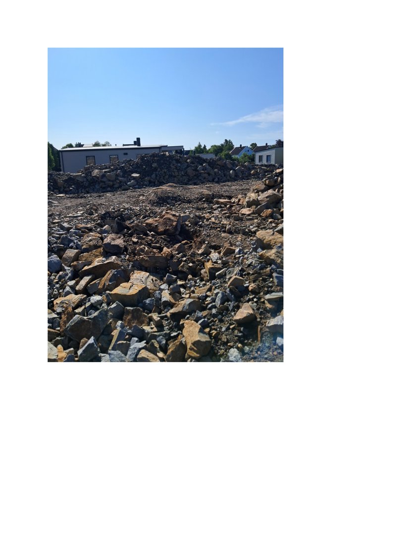 Fotografi av en byggeplass eller tomt med stein og grus i forgrunnen, og en bygning i bakgrunnen.
