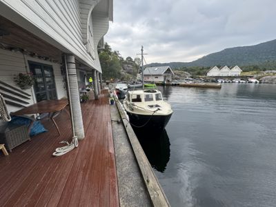 Fotografi av en villa med terrasse og brygge ved vannet, med båter og andre hus i bakgrunnen.