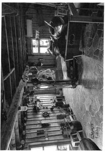 Sort-hvitt fotografi av et interiør i en bygning, sannsynligvis et garasje eller verksted, som viser en bil, verktøy og utstyr.