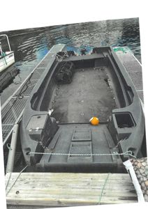 Fotografi av en liten båt (kanskje en fiskebåt eller kutter) som er festet til en kai eller ponton. Båten er tom for innredning og har en motor montert på bakhellet.