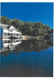 Fotografi av en bygning ved vannet med båter og skog i bakgrunnen.