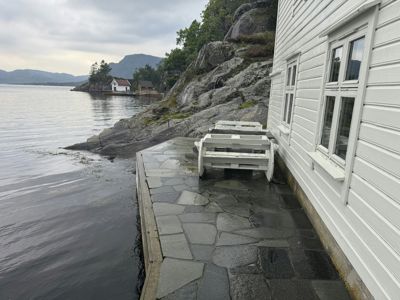 Fotografi av en hvit, trepanelert bygning med en steinlagt terrasse og en hvit båt som ligger på terrassen. I bakgrunnen ses en kystlinje med fjell og andre hus.