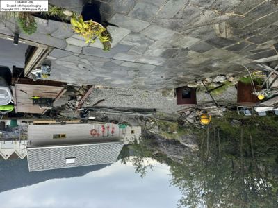 Fotografi av en bygning sett fra siden, med en steinlagt terrasse, en liten uthus eller båthavn, og en bakke med trær i bakgrunnen.