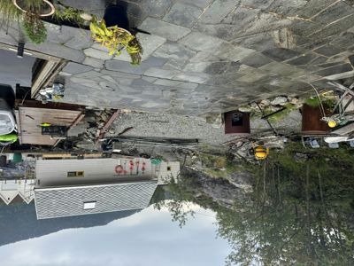 Fotografi av en bygning med grå takfliser, en steinlagt terrasse med planter, og en bakgrunn med fjell og trær.