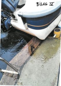 Fotografi av en båt som ligger i vann ved en kai eller brygge. Bildet viser detaljer som motor, båtens skrog, en trebjelke i vannet og regndråper.