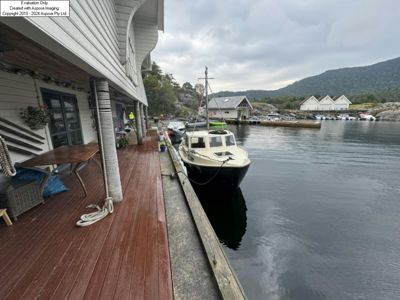 Fotografi av en hytte med uteplass ved en kai, med båter og fjell i bakgrunnen.