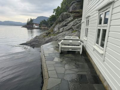 Fotografi av en hvit, panelt husfasade ved kanten av en steinete kyst. Bildet viser en steinlagt terrasse med en hvit benk, og i bakgrunnen er det vann, fjell og andre hus.