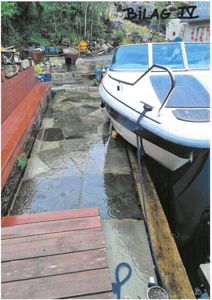 Fotografi av en brygge med en båt, en steinlagt gangvei og en del av en bygning i bakgrunnen.