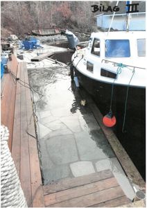 Fotografi av en brygge med en båt ved kai. I bakgrunnen er det en annen båt og en bygning.