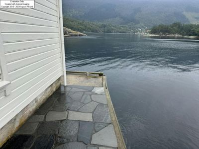 Fotografi av en bygning med hvit panelvegg og en steinbelagt terrasse som går ut over vannet, med fjell og skog i bakgrunnen.