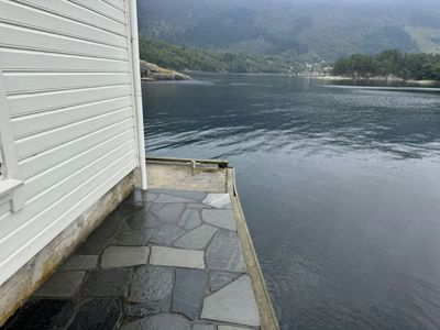 Fotografi av en bygning med hvit panelvegg og en steinbelagt terrasse som vender ut mot vann og fjell.