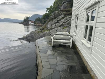 Fotografi av en hvit, trepanelert bygning ved kysten med en steinlagt terrasse og en båtstige. I bakgrunnen ses fjell og andre hus.