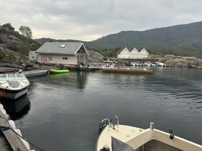 Fotografi av en bygning ved vannet med båter og en fjellbakgrunn.