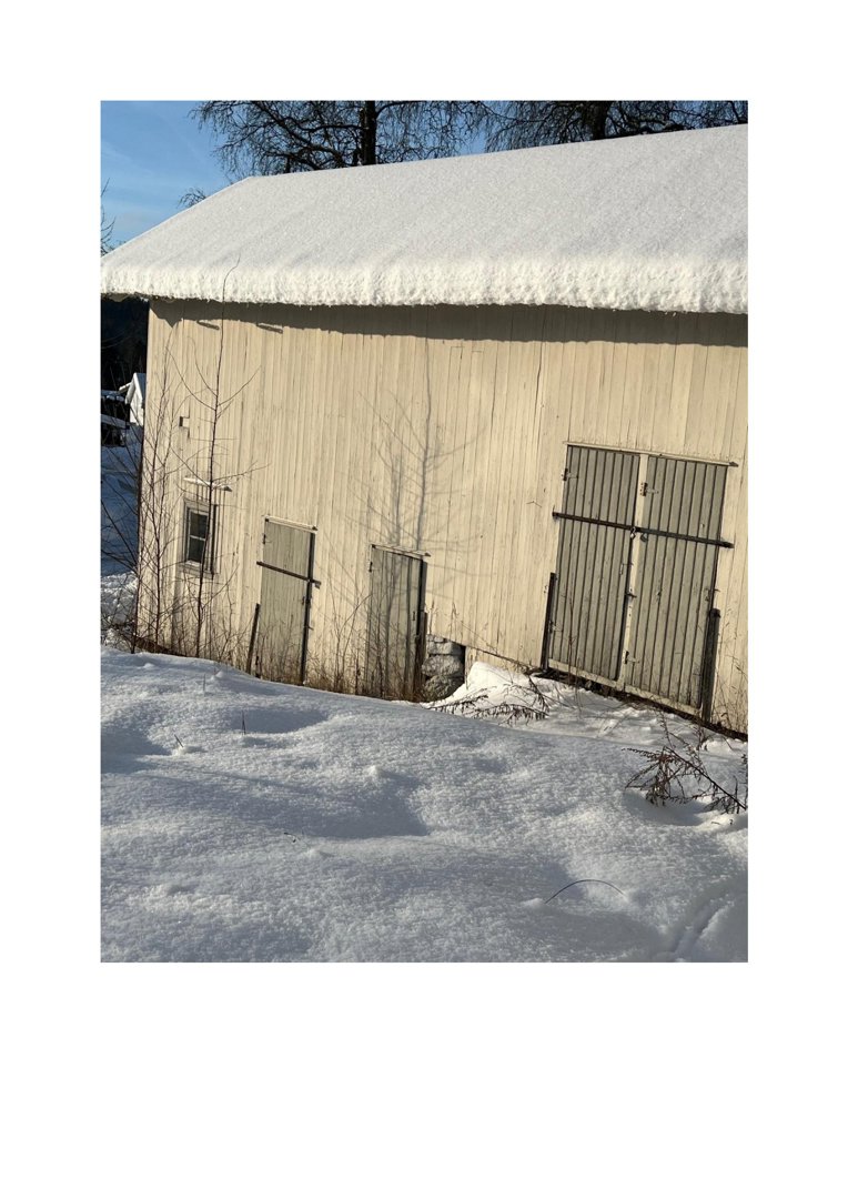 Fotografi av en hvit, trepanelert bygning (sannsynligvis en uthus eller garasje) med snø på tak og på bakken.