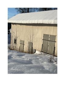 Fotografi av en hvit, trepanelert bygning (sannsynligvis en uthus eller garasje) med snø på tak og på bakken.