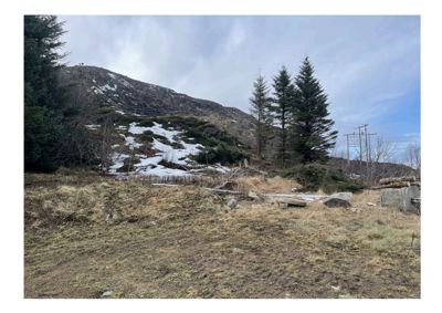 Fotografi av et tomt terreng med en skråning, trær og snøflekker i bakgrunnen.