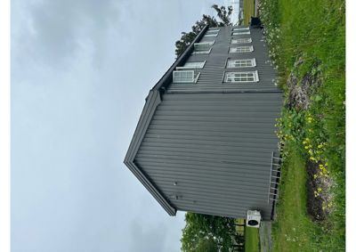 Fotografi av en grå, trehusliknende bygning sett fra siden. Bygningen har flere vinduer og er plassert på en grønn tomt med gress og urter.