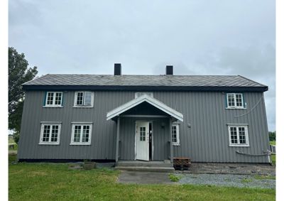 Fotografi av en grå, toetasjes bygning sett fra fronten. Bygningen har et tak med to røykkanaler, vinduer på begge etasjer og en liten portoverbygning foran hoveddøren. Bildet viser fasaden og umiddelbar omgivelse.