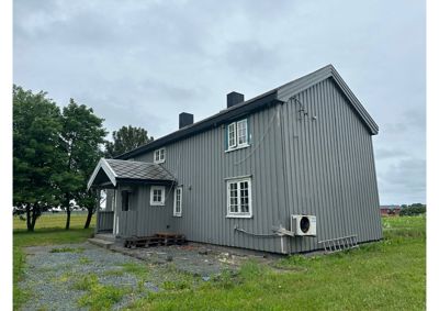 Fotografi av en grå, trepanelt bygning sett fra siden. Bildet viser fasaden, vinduer, tak, en liten veranda og omgivelsene med gress og trær.
