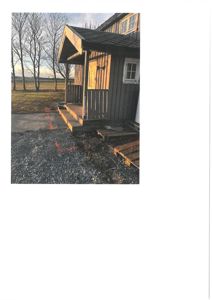 Fotografi av en liten, mørk bygning med et forhøyet inngangsparti og en veranda. Bildet viser fasaden og omgivelsene, inkludert grus og barne trær.