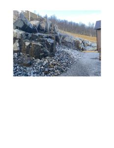 Fotografi av en utsmykket steinvegg med en liten vannfall, en grusvei og en del av en bygning i høyre hjørne.