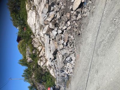 Fotografi av en steinmur eller bergvegg ved en vei, med en del av en bygning i bakgrunnen.