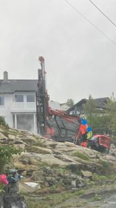 Fotografi av en byggeplass med boremaskin og arbeidere ved en hvit bolig.