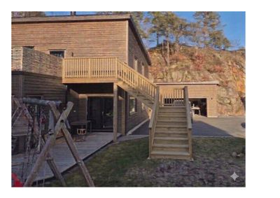 Fotografi av en ferdigbygd hytte med utvendig trapp, balkong og garasje sett fra bak/side.