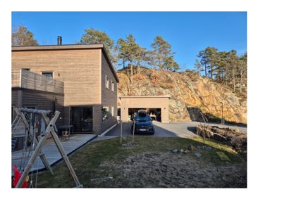 Fotografi av en moderne, trepanelt bolig med garasje og uteplass, sett fra bakgården mot en steil fjellside.