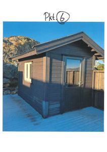 Fotografi av en mørk, trepanelert hytte eller uthus med etasjehus, sett fra en vinkel som viser både fasade og sidevegg. Bygningen står på en dekket terrasse med en fjellside i bakgrunnen.