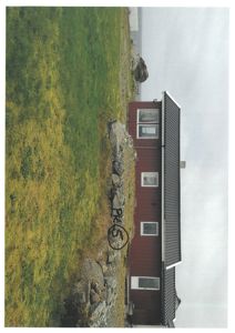 Fotografi av en rød, toetasjes hytte eller hus sett fra siden, med grønt gress og steiner i forgrunnen.