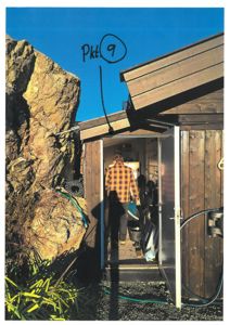 Fotografi av en liten, mørk trehytte eller uthus med en person som går inn i døren. Bildet viser bygningens utside, en stor steinblokk til venstre og en blå himmel.