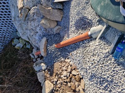 Fotografi av en utendørs byggeplass eller grunnsituasjon med stein, grus, en oransje rørledning og en vannflaske.