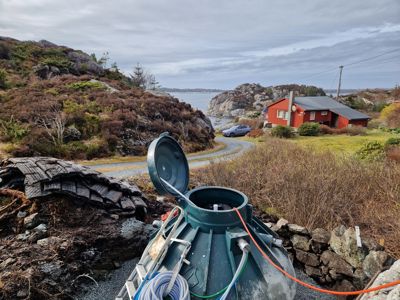 Fotografi av en byggeplass eller tomt med et grønt septiktank i forgrunnen, og en rød hytte i bakgrunnen.