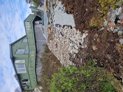 Fotografi av en grønn hytte sett fra siden, med en steinmur og grus i forgrunnen.