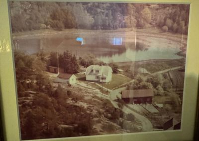 Et gammelt fotografi av en eiendom med flere bygninger ved en vannflate, sett fra et høydepunkt.