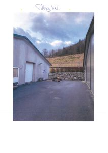 Fotografi av en bygning sett fra utsiden, med en asfaltert flate i forgrunnen og en steinmur i bakgrunnen.