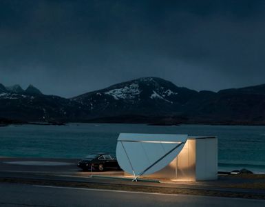 Fotografi av en moderne, hvit bygning med et karakteristisk, skjeformet tak sett fra utsiden mot havet og fjellene.