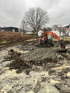 Fotografi av en byggeplass med en oransje gravemaskin (Kobelco) som jobber med å grave i en mudderete grøft. I bakgrunnen ser man nabolagshus og trær.