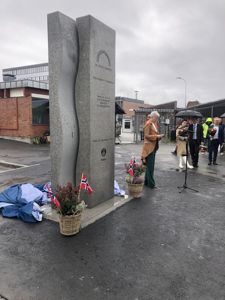 Fotografi av en minnestøtte eller monument med personer og flagg i bakgrunnen.